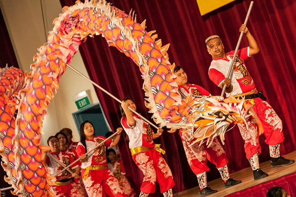 Performers in red costumes hold poles to maneuver a large orange dragon prop on a stage with red curtains.